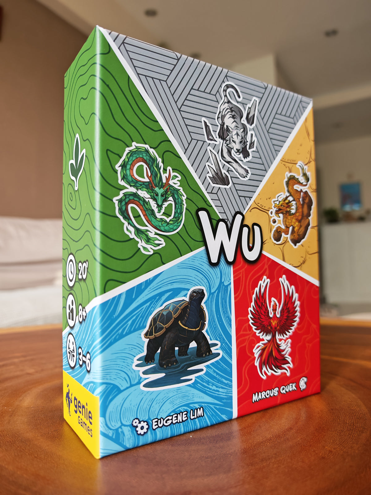 The product box for the board game 'Wu' by Eugene Lim and Marcus Quek sits on a wooden table in a blurred living room setting. The box features a colorful, geometric design with four quadrants representing the Chinese WuXing elements: a green dragon, a white tiger, a yellow dragon, a red phoenix, and a black tortoise, with a large white 'Wu' logo in the center.