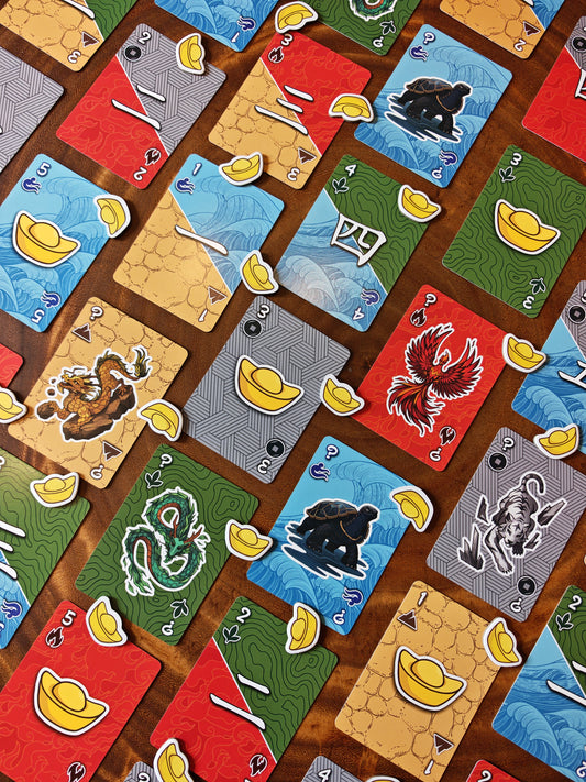 A close-up overhead view of a card game board with multiple colorful cards arranged on a wooden surface. The cards feature illustrated symbols including yellow ingots, a red phoenix, a green dragon, black turtle, and other decorative graphics.